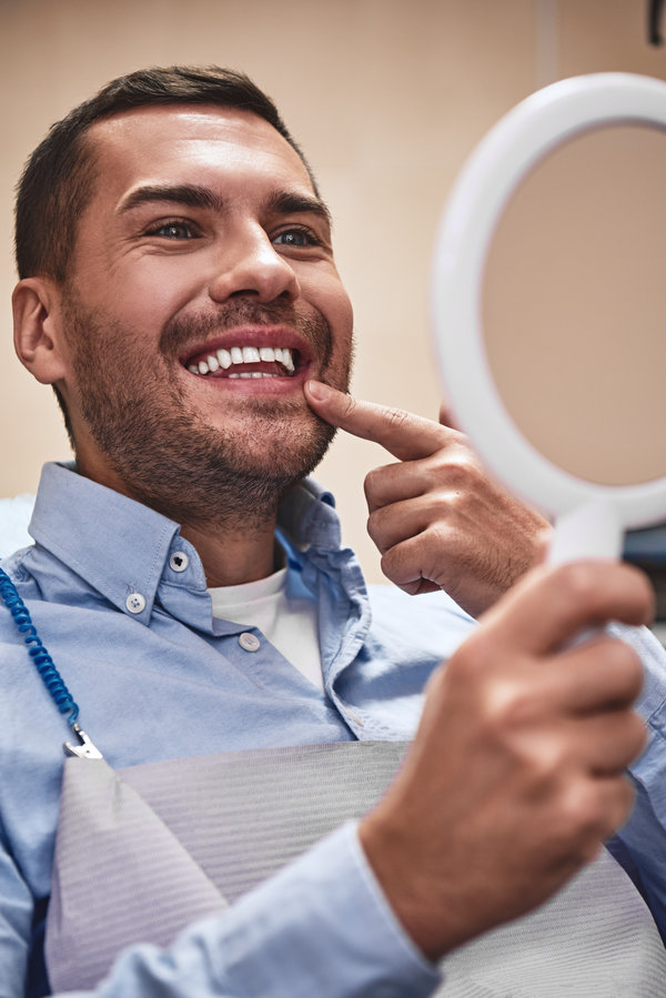 Cheerful man in casual wear looking in the mirror while sitting in the dental chair after treatment. He is satisfied with the result. Modern dental clinic. Vertical shot