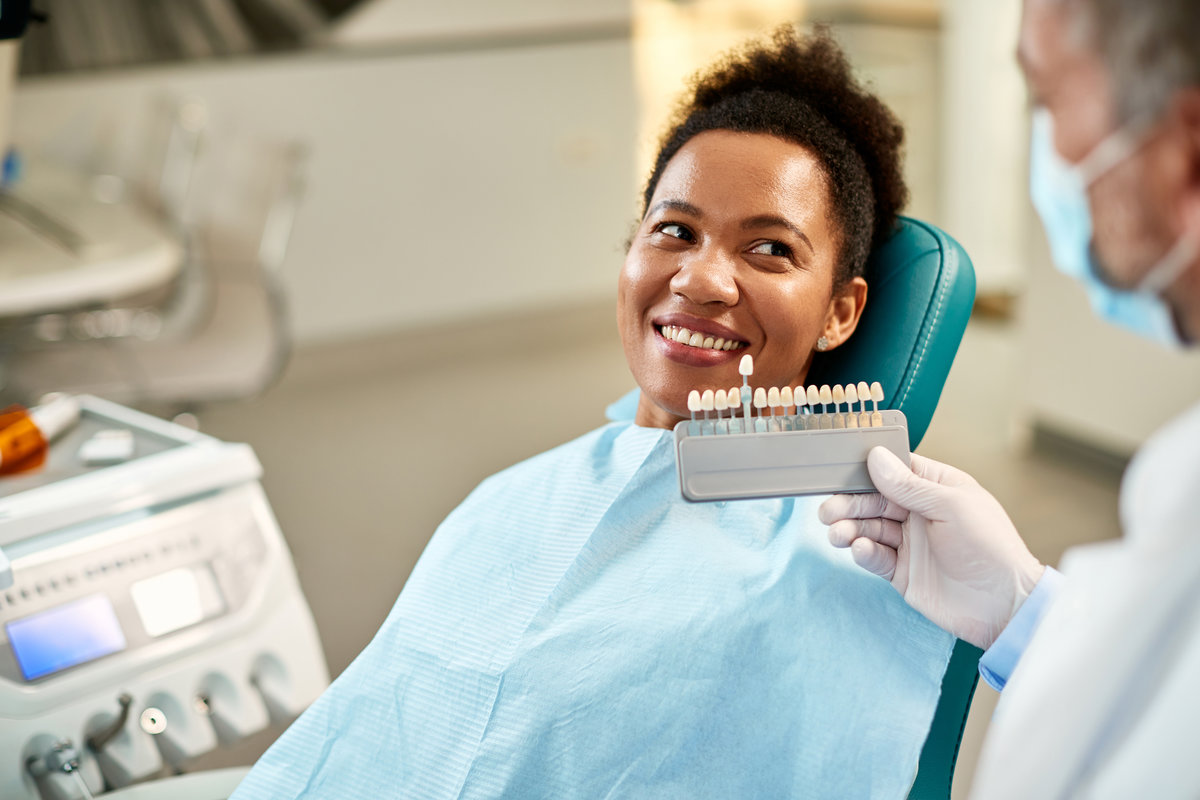 Happy black woman and her dentist choosing shade of dental veneers during appointment at dentist's office.