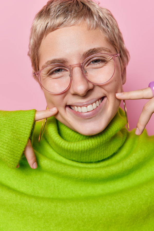 Headshot of positive adult woman with short blonde hair points at mouth shows white perfect teeth wears big transparent eyeglasses and warm green jumper with collar isolated over pink background