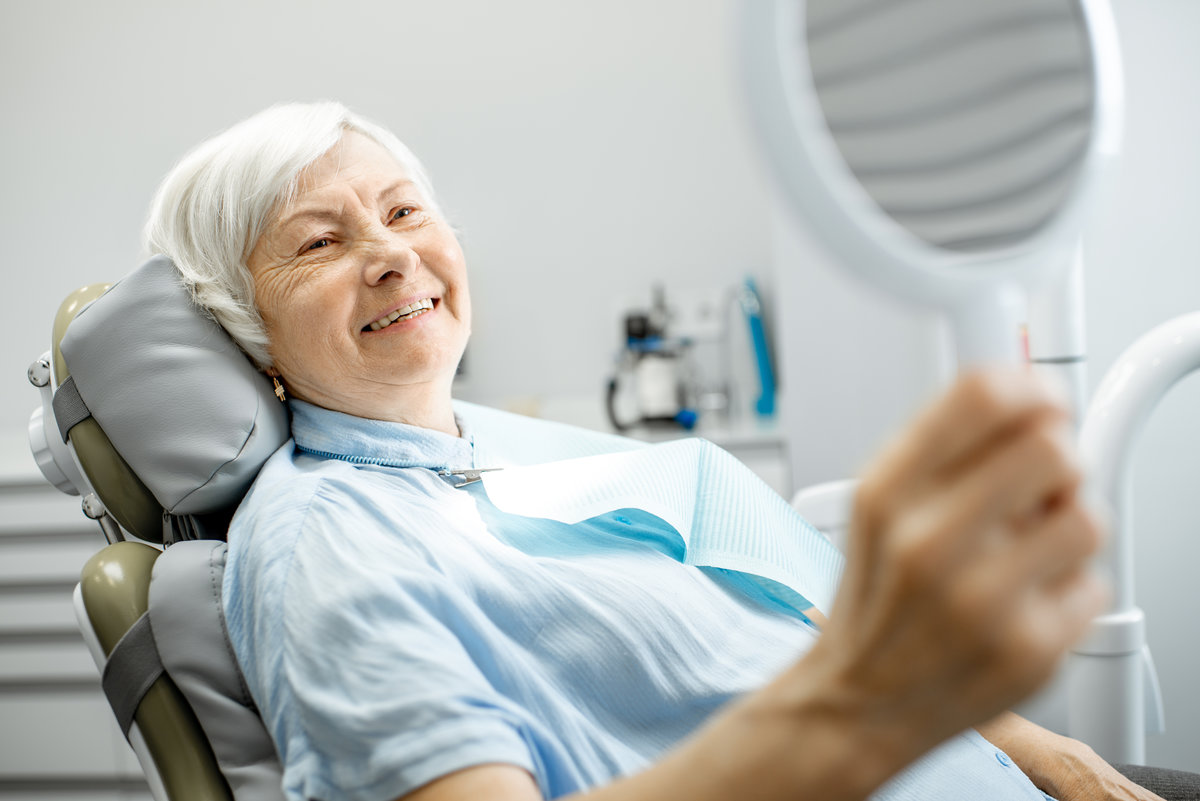 Happy elderly woman enjoying her beautiful toothy smile looking to the mirror in the dental office