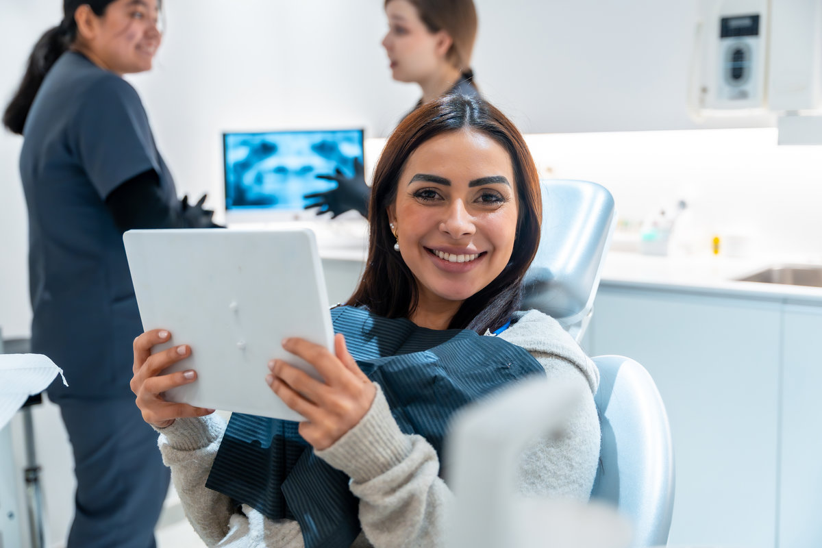 Smiling woman holding a mirror in a dentist chair, expressing satisfaction with dental results while staff discusses radiography