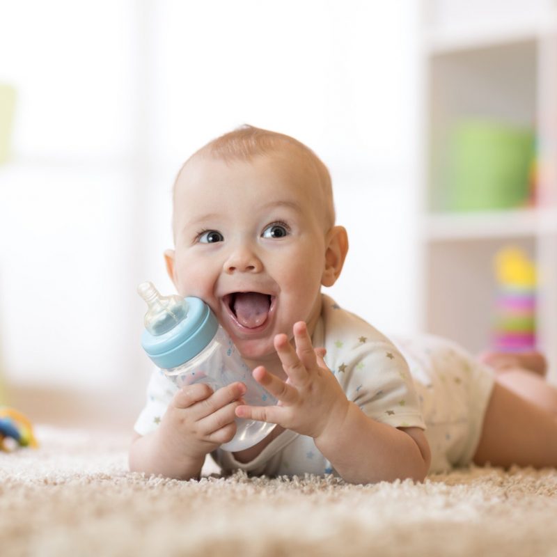 Cute baby boy drinking from bottle. Kid lying on carpet in nursery at home. Smiling child is 7 months old.